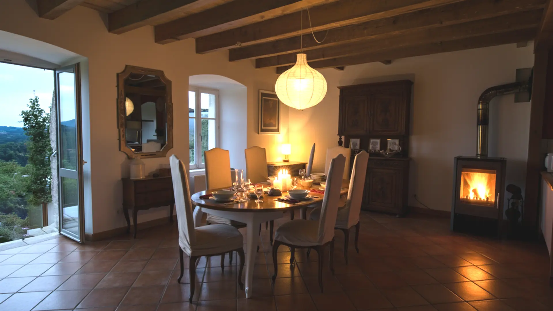 Salle à manger chaleureuse éclairée à la lueur des bougies et du feu de bois. Une grande table ronde est dressée pour un dîner, avec assiettes, verres et serviettes soigneusement disposés. Les chaises tapissées entourent la table, baignées dans une lumière douce venue d’une lampe de table et d’un grand lampion suspendu. Les poutres en bois au plafond donnent une ambiance rustique et apaisante. À gauche, une porte-fenêtre ouverte laisse entrevoir le paysage du soir et les collines verdoyantes. L’ensemble dégage une atmosphère intime et confortable, parfaite pour un repas tranquille après une journée dans la nature.