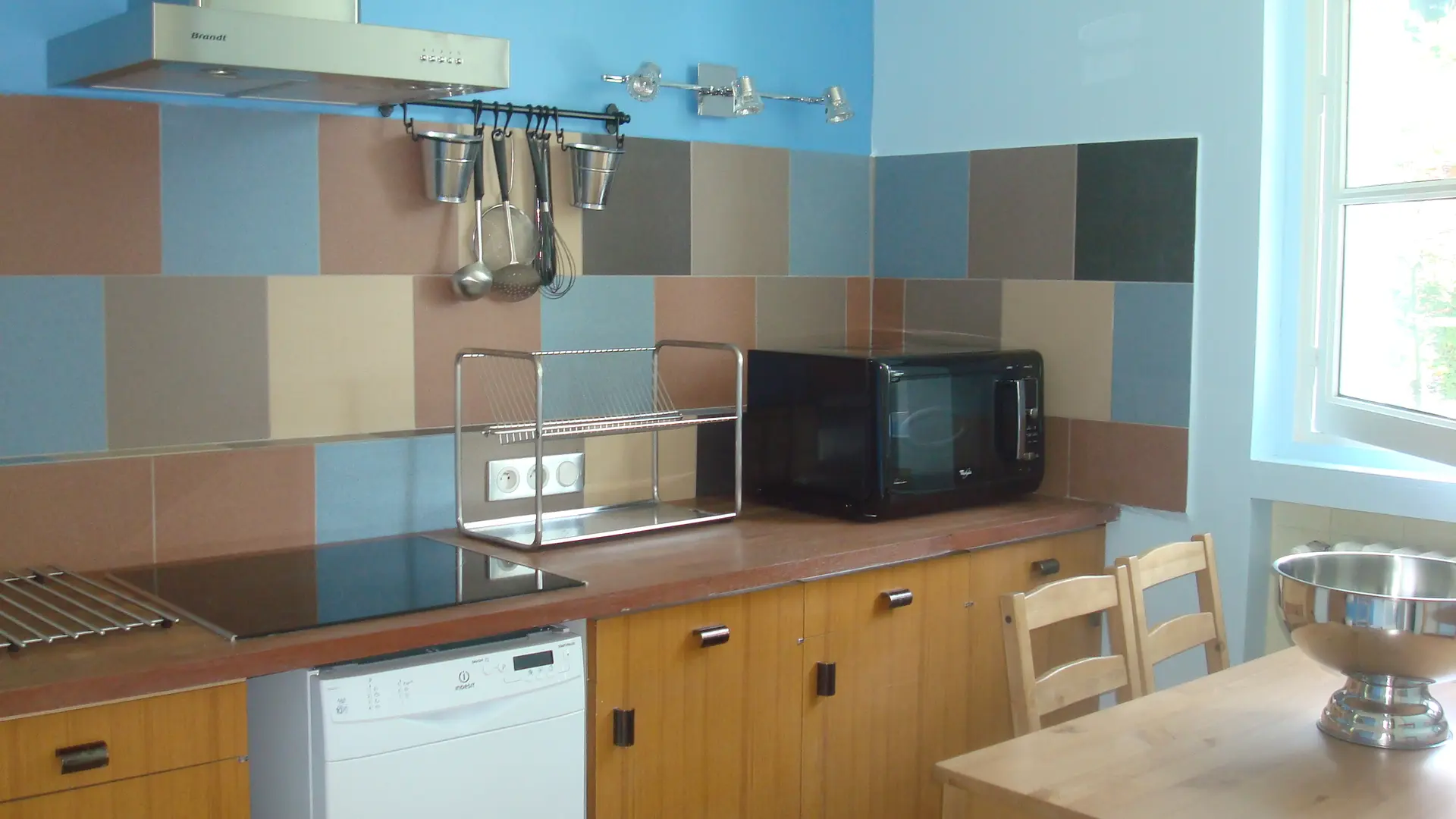 Cette cuisine est équipée d'une plaque de cuisson, d'un four micro-ondes et d'un réfrigérateur. Les murs sont recouverts de carreaux de faïence de différentes couleurs et les meubles sont en bois clair. Une table et des chaises en bois complètent l'aménagement.