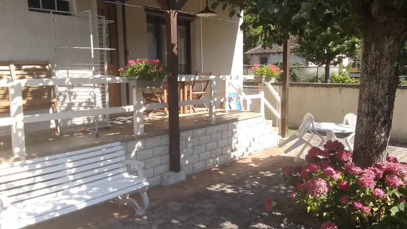 La façade d'une maison avec une terrasse aménagée de mobilier de jardin et un arbre sur la droite au premier plan, devant des massifs de fleurs.