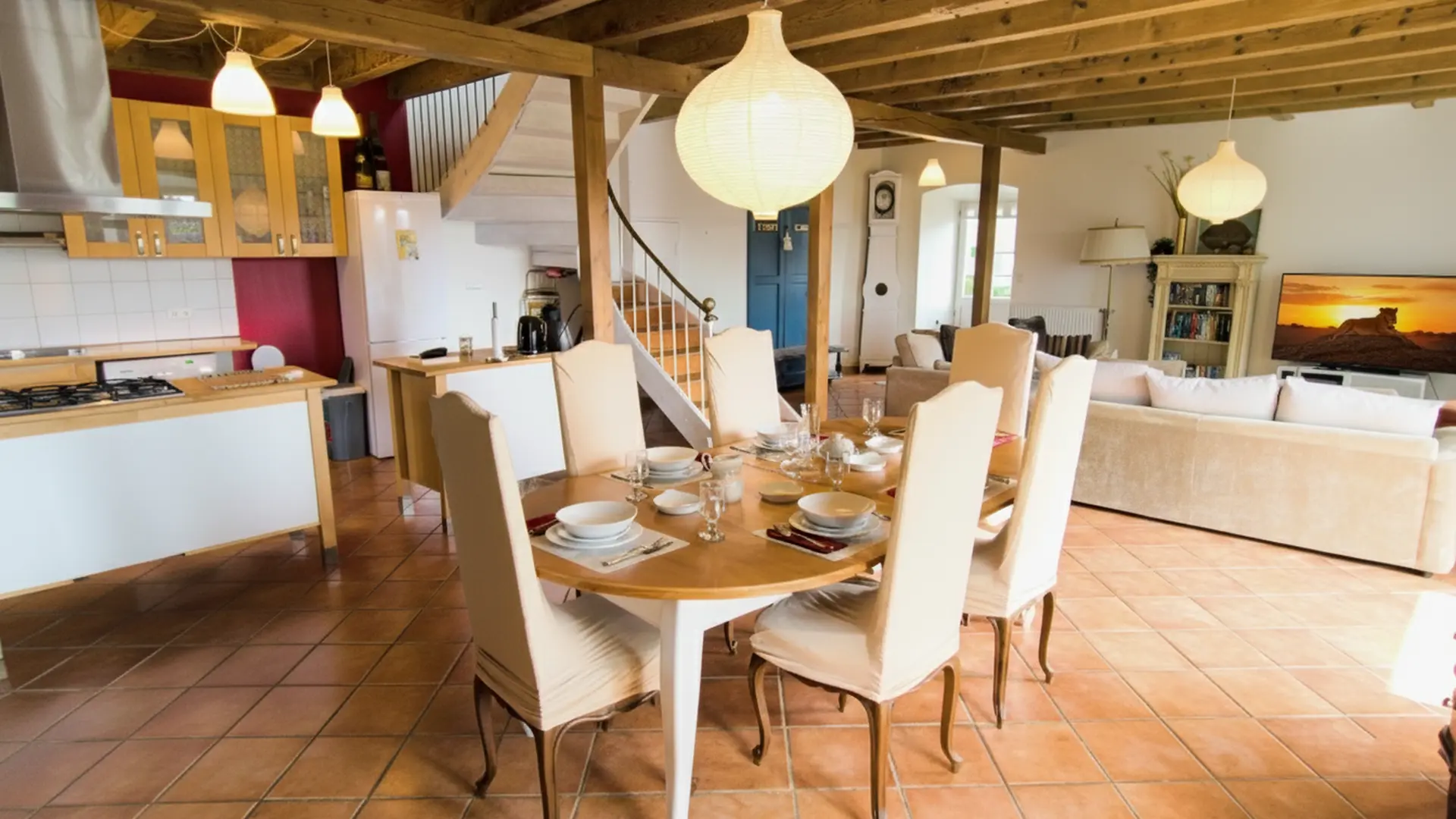 Une salle à manger et une cuisine ouvertes avec un sol en carreaux de terre cuite. Au centre, une table ronde en bois est dressée avec six chaises à dossier haut et des sets de table beiges. Au-dessus de la table, un grand luminaire sphérique blanc est suspendu au plafond en bois avec des poutres apparentes. À l'arrière-plan, à gauche, se trouve une cuisine moderne avec des armoires en bois clair, un îlot de cuisine blanc et un mur d'accent rouge foncé. À l'arrière-plan, à droite, on aperçoit un salon avec un canapé beige, une télévision affichant un paysage désertique, et une bibliothèque blanche remplie de livres. Un escalier  en bois mène à l'étage supérieur.