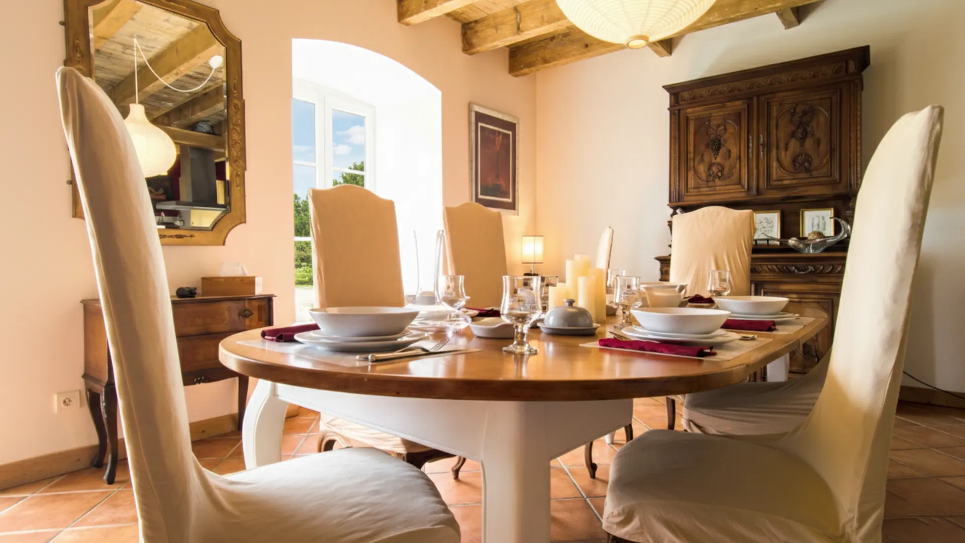 Un gros plan d'une table de salle à manger en bois ovale dressée avec de la vaisselle, des bougies et des couverts. La table est entourée de chaises à housses beiges. À gauche, un grand miroir doré est accroché au-dessus d'une petite commode en bois sombre, avec une fenêtre derrière elle. À droite, un grand buffet en bois sculpté sombre se dresse contre le mur. La lumière du jour éclaire la pièce par la fenêtre et une ambiance chaleureuse est créée par un luminaire suspendu visible en haut.