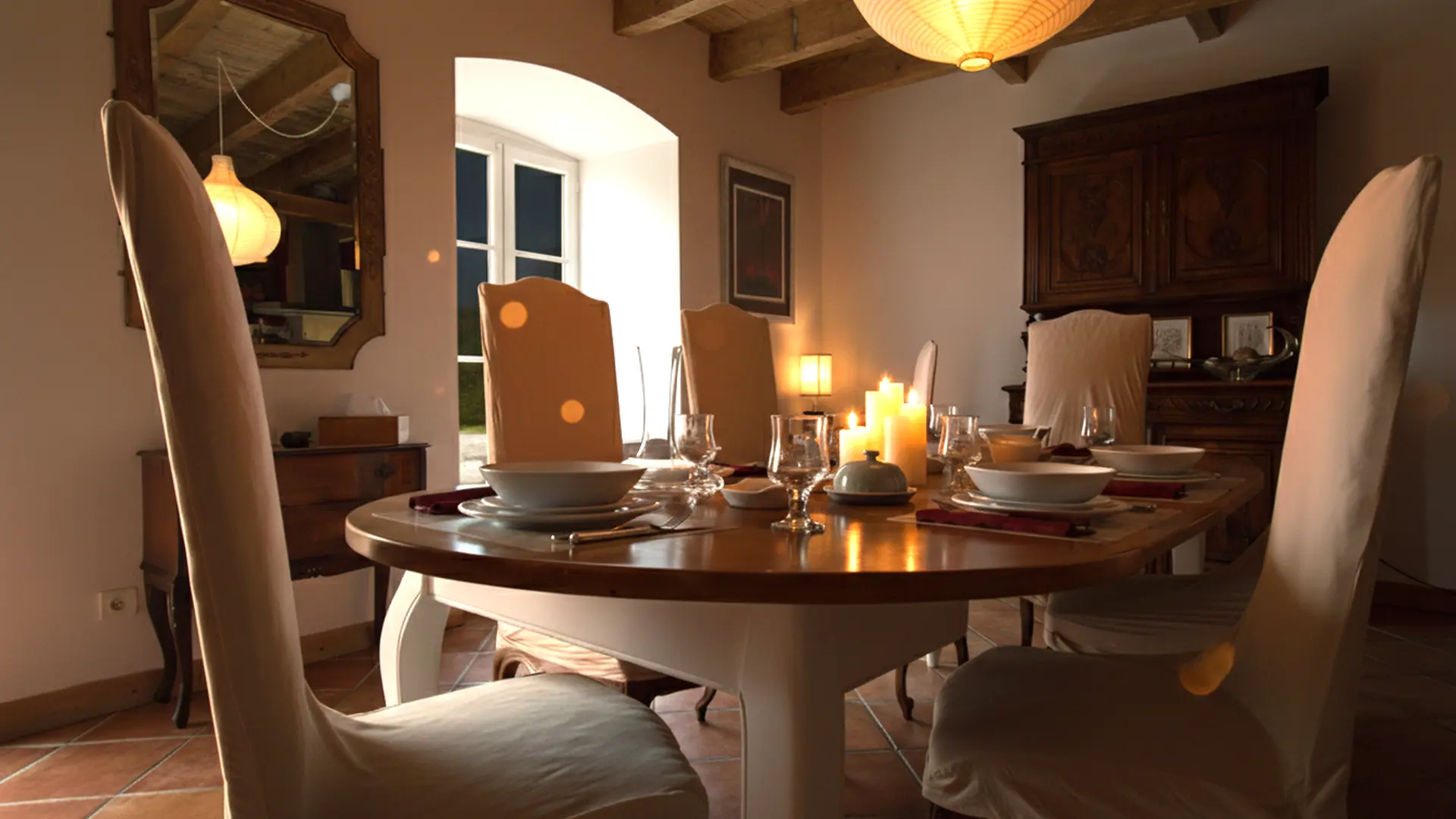 Une salle à manger chaleureuse et élégante, vue sous un angle rapproché, photographiée de nuit. Une table ovale en bois est dressée au centre avec de la vaisselle blanche, des couverts et des bougies allumées qui diffusent une lumière douce. La table est entourée de chaises à dossier haut recouvertes de housses beiges. À gauche, un grand miroir doré est accroché au mur au-dessus d'une petite commode en bois foncé. Une fenêtre arquée à carreaux est visible à l'arrière-plan, laissant entrer une lumière extérieure légèrement bleutée, ce qui confirme la prise de vue nocturne. À droite, un grand buffet ancien en bois sombre, richement sculpté, se dresse contre le mur. Des poutres en bois apparentes traversent le plafond. La pièce est éclairée par la lumière des bougies et d'un luminaire suspendu sphérique en haut de l'image.