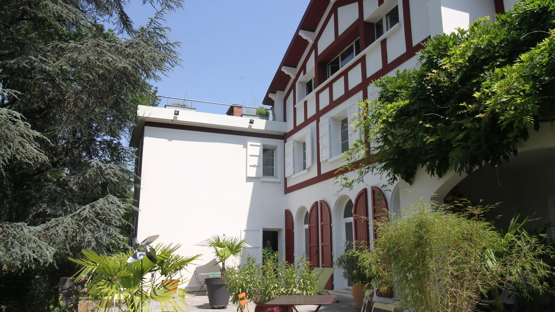 Cette façade est constituée d'un bâtiment à colombages, avec des murs blancs et des poutres en bois apparentes de couleur rouge. Un jardin arboré se trouve devant le bâtiment.