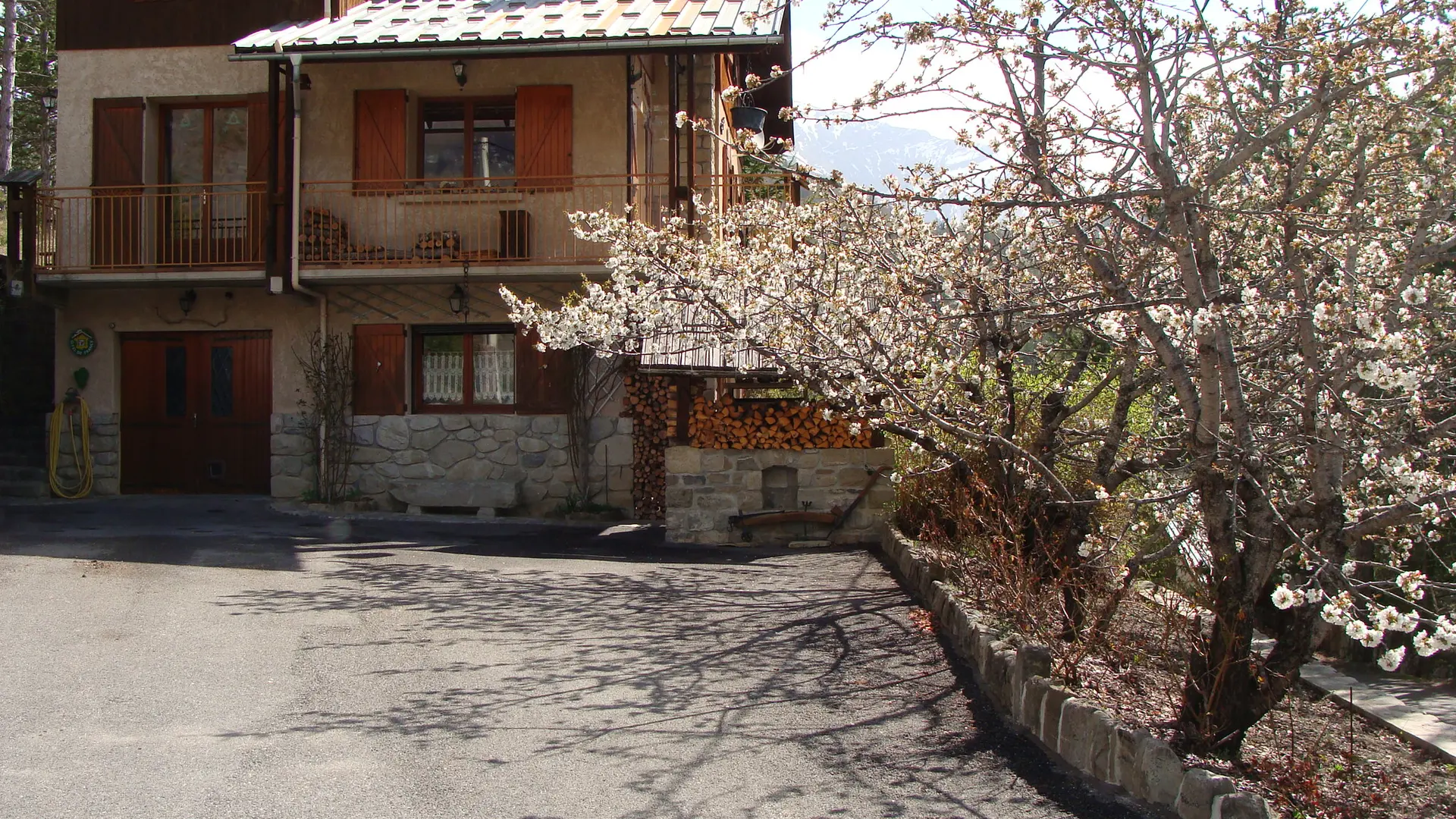 Vue de face de maison en pierre et bois sur trois étages, cour intérieure bitumée, arbre en fleurs