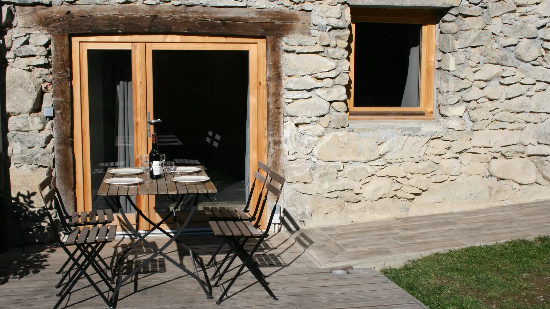 Des portes vitrées donnent sur une petite terrasse devant un mur en pierre d'une maison. Une table est dressée pour 4 personnes.