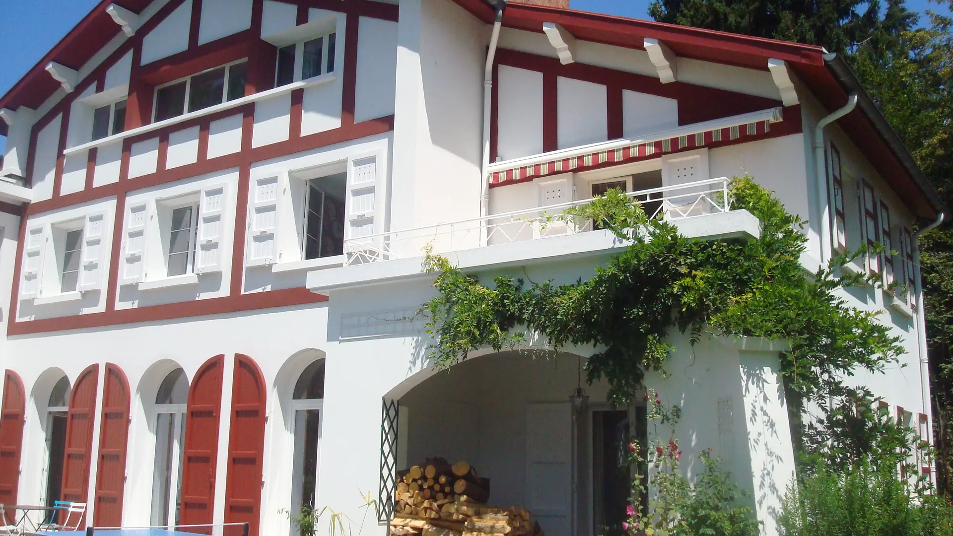 Cette maison de style thermal présente une façade à colombage avec des murs blancs et des poutres en bois rouge. Les fenêtres sont larges et équipées de volets battants rouges. Le toit est pentu et recouvert de tuiles.