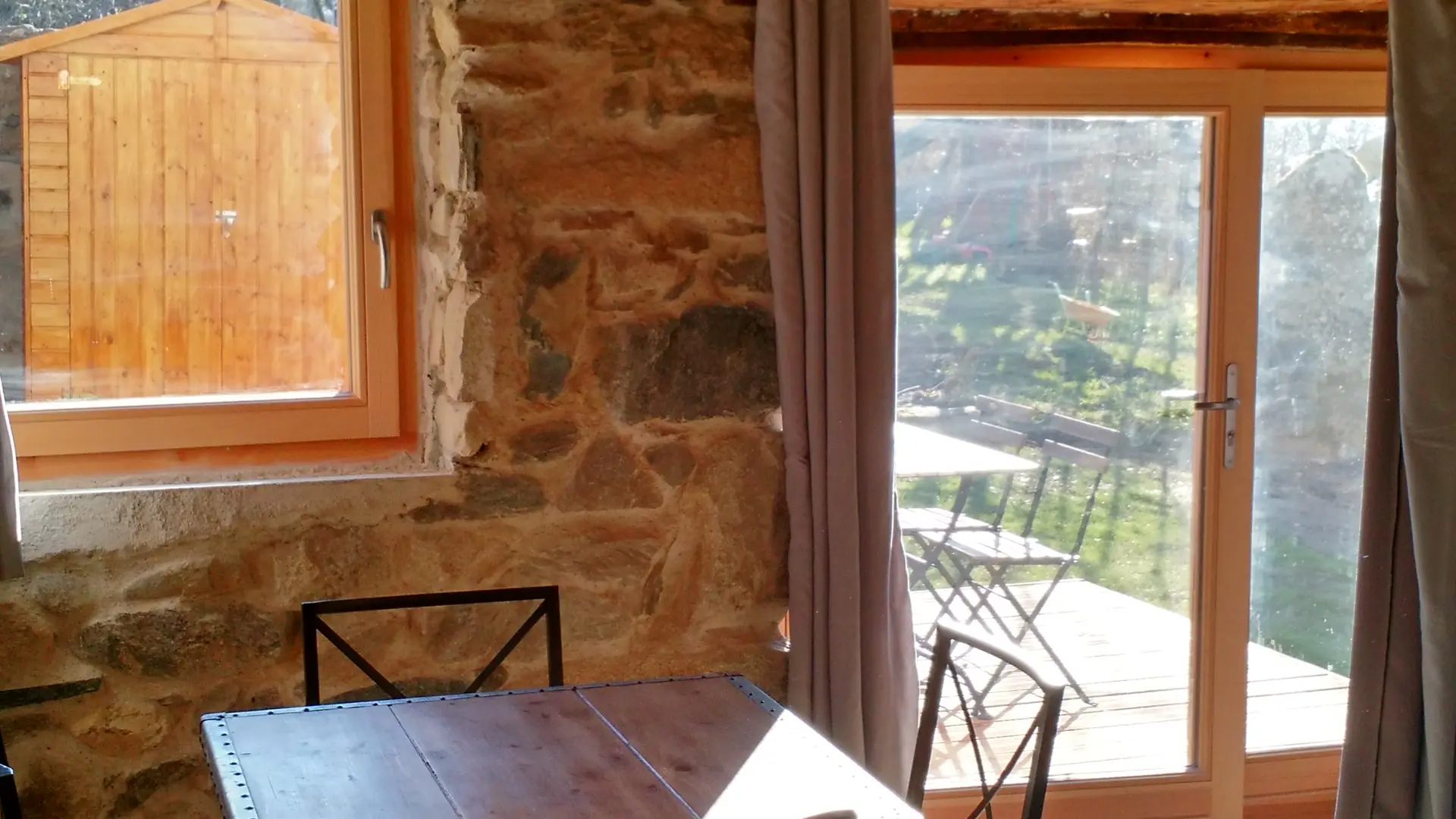 Une pièce chaleureuse avec un plafond en bois et un mur en pierre, meublée d'une table et d'un canapé, s'ouvre sur une terrasse extérieure.