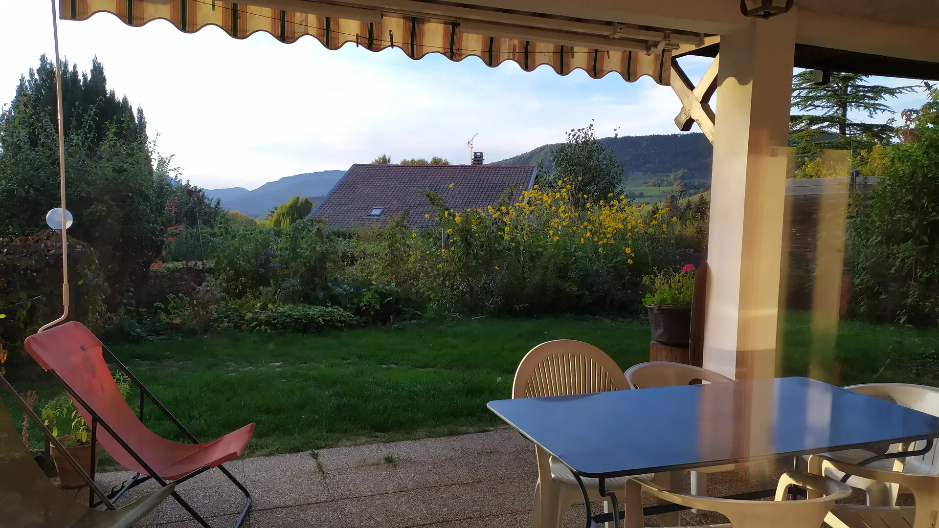 La terrasse est aménagée avec une table, des chaises et un fauteuil, et couverte d'un auvent rayé. La vue est dégagée sur un jardin verdoyant et des collines en arrière-plan.