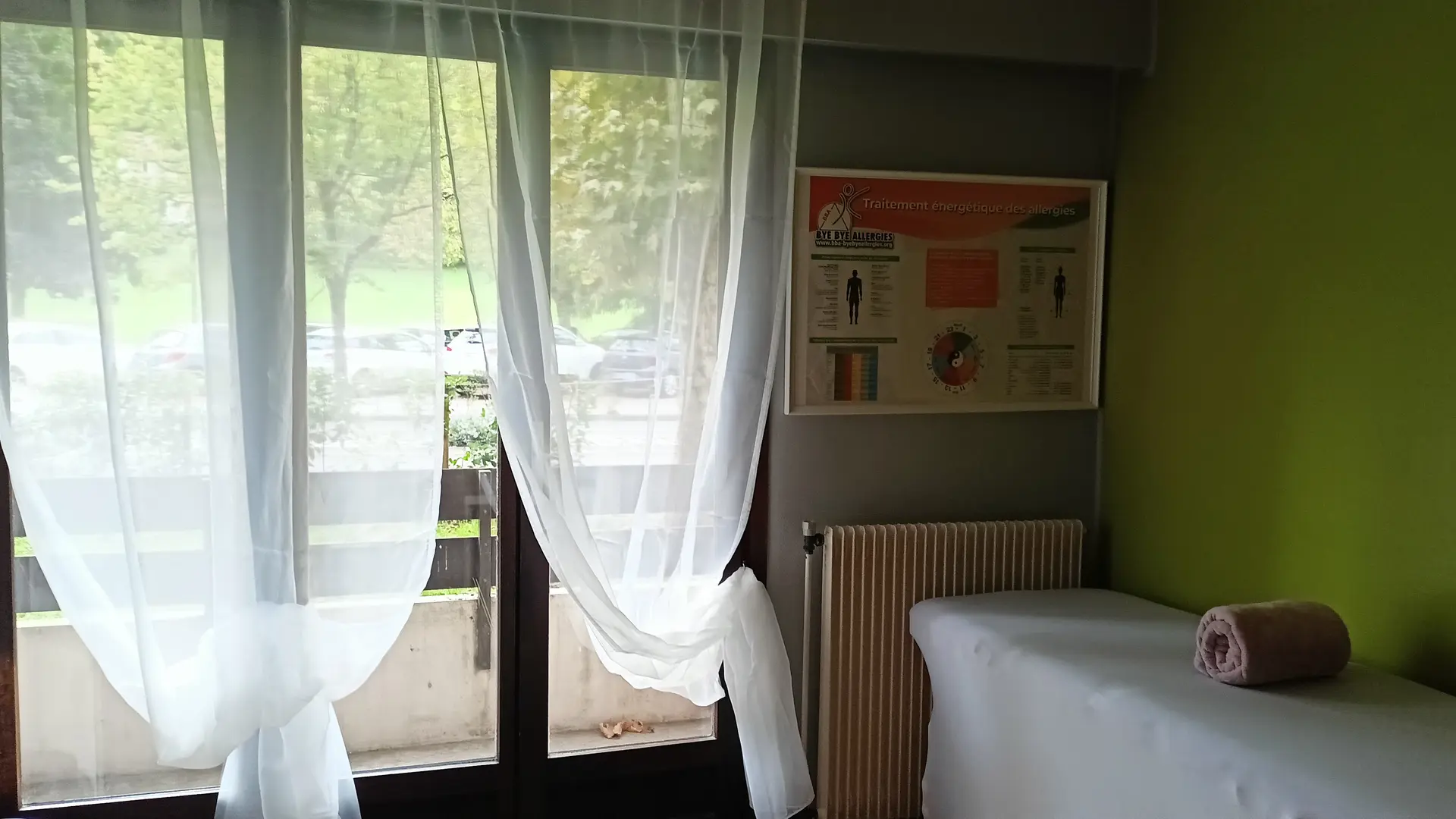 C'est une pièce lumineuse avec un mur vert, une fenêtre donnant sur des arbres et une rue avec des voitures garées, et une table de massage recouverte d'un drap blanc avec une serviette enroulée.