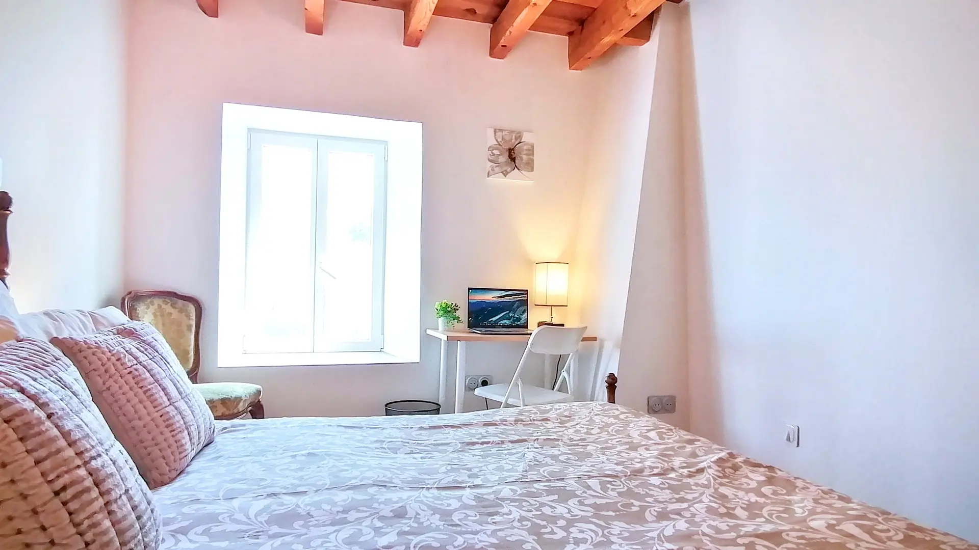 C'est une chambre lumineuse aux couleurs douces (blanc, vert pastel, beige). Le plafond comporte des poutres, les murs sont blancs. Le lit deux personnes est aussi en bois ainsi que la tête de lit. De chaque côté, on trouve des tables de nuit en bois avec une lampe de chevet, ainsi qu'un fauteuil près de la fenêtre et un bureau de l'autre côté avec écran et poubelle à droite du bureau. Les deux pans de murs sont décorés par des tableaux avec des  motifs de végétaux.