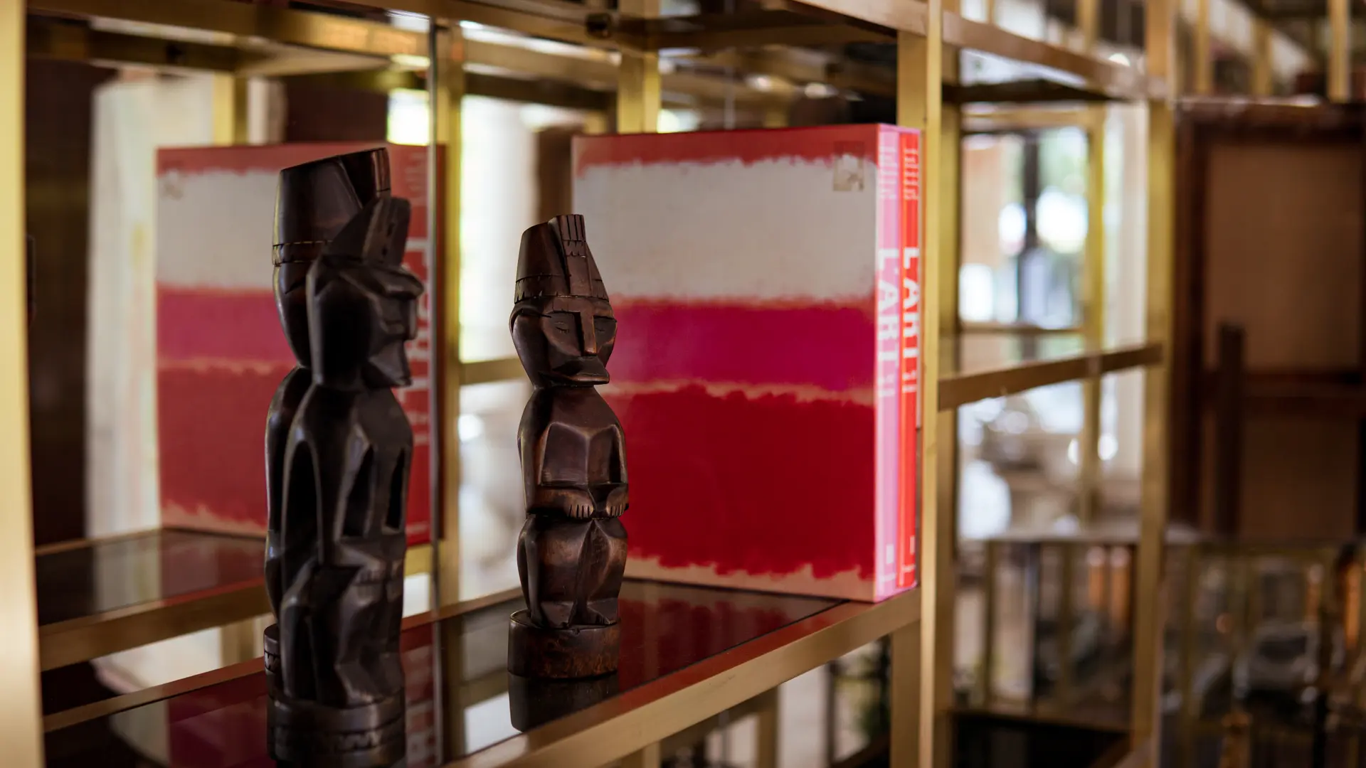 Deux statuettes en bois se détachent sur un fond de rayonnages dorés où reposent livres et objets d’art.