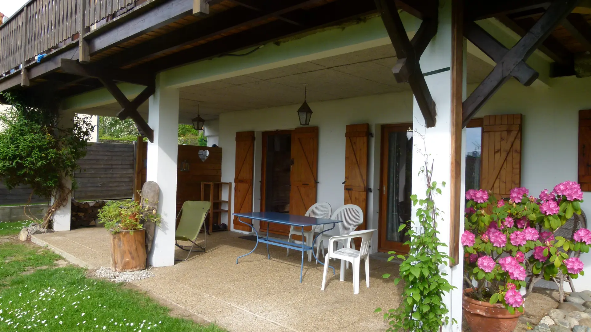 Une terrasse en rez-de-jardin sous un balcon en bois avec une table et des chaises de jardin.