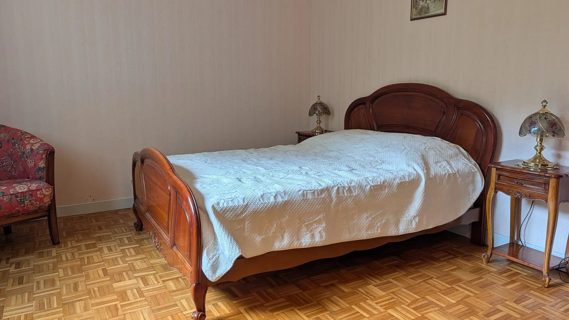 Une chambre à coucher avec des murs unis et un parquet en bois. On y trouve un lit double en bois sculpté avec un couvre-lit bleu, une table de chevet avec une lampe à droite, et un fauteuil en tissu rouge-marron à gauche.
