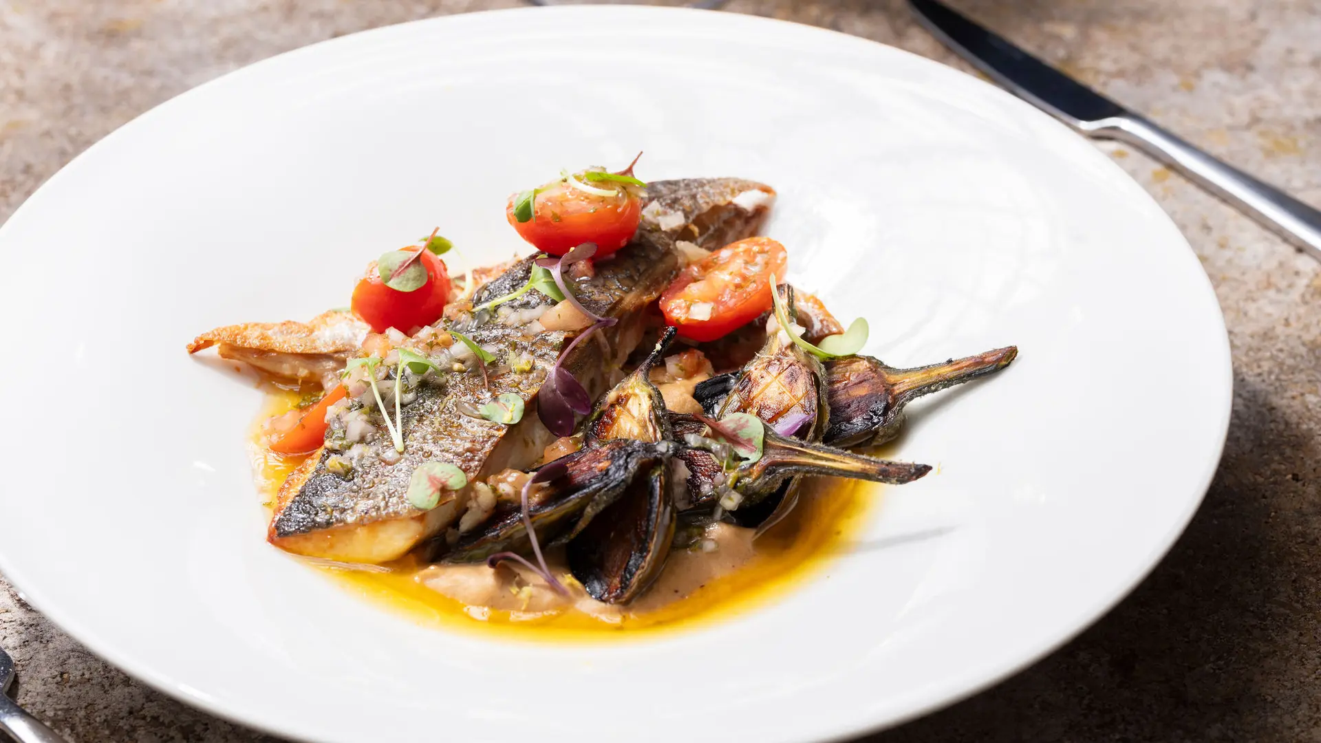 Cette assiette présente un poisson cuit, avec une peau croustillante et une chair blanche et feuilletée, accompagné de légumes méditerranéens comme des artichauds et des tomates cerises, le tout nappé d'une sauce jaune.