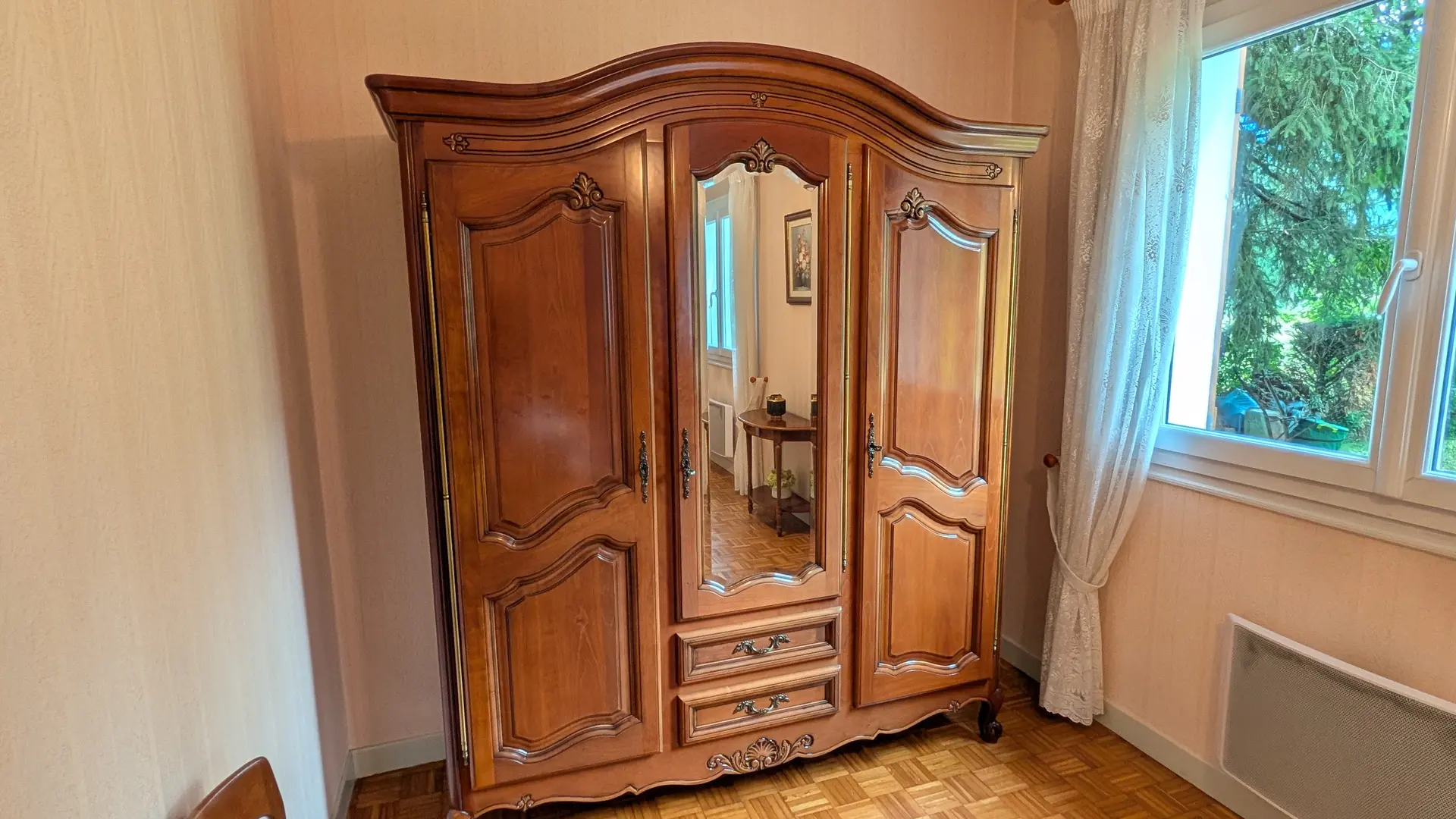 Une grande armoire en bois de style traditionnel avec une porte centrale en miroir est positionnée contre un mur dans une pièce au parquet en bois.