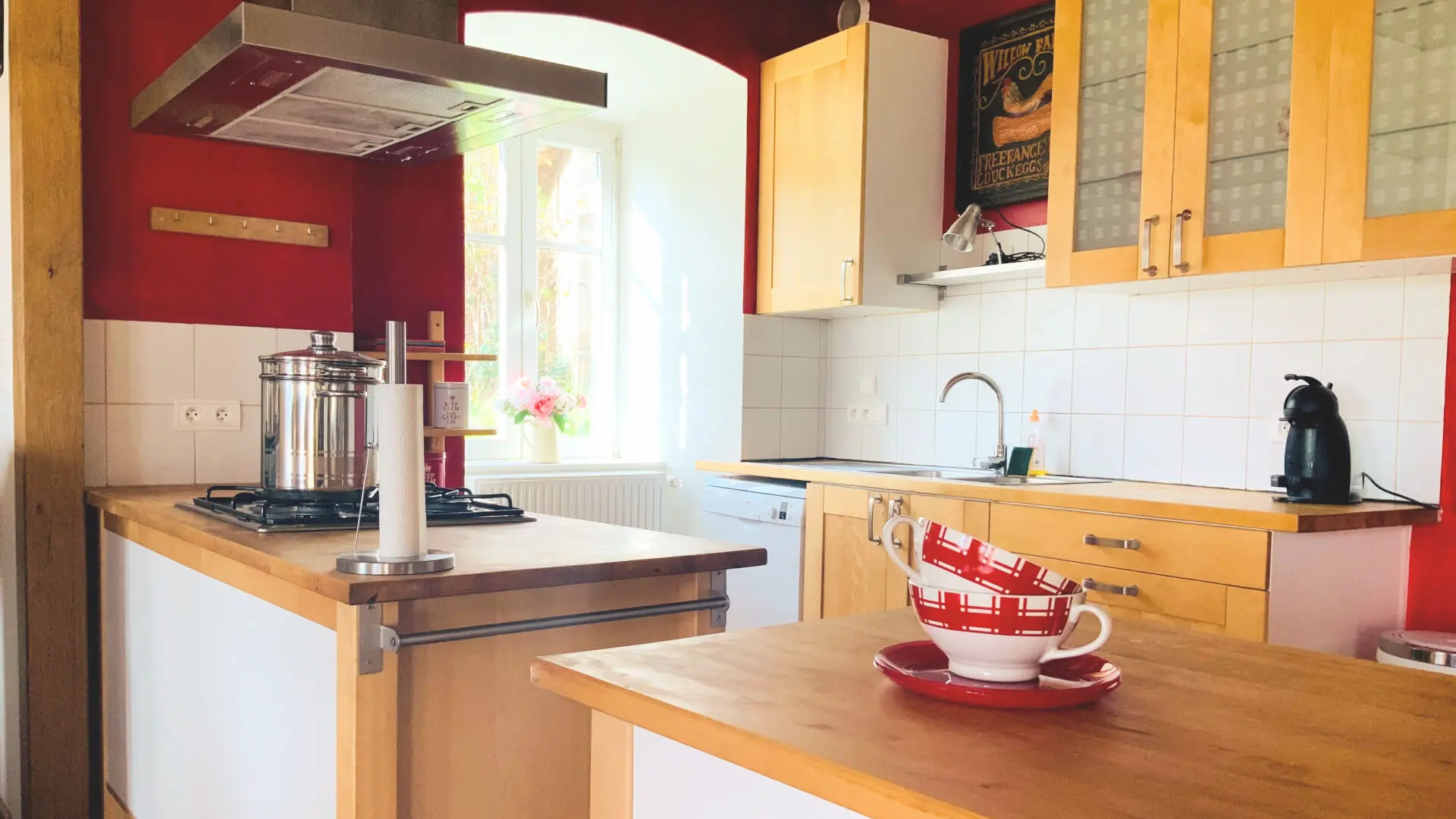 Une cuisine ouverte sur la salle à manger est très fonctionnelle avec des meubles en bois, une hotte au-dessus de la table de cuisson. A l'opposé, un évier avec le lave-vaisselle et sur la table de travail, il y a une Senseo. Une grande fenêtre  apporte beaucoup de lumière avec pour décoration un pot de fleurs. Un couscoussier vous attend pour préparer de bons plats, Sur l'autre table de travail, il y a deux bols blancs et carreaux rouges et deux assiettes à dessert rouges. Le premier tiers des murs est recouvert de carrelage blanc, le reste des murs est de couleur terracotta.