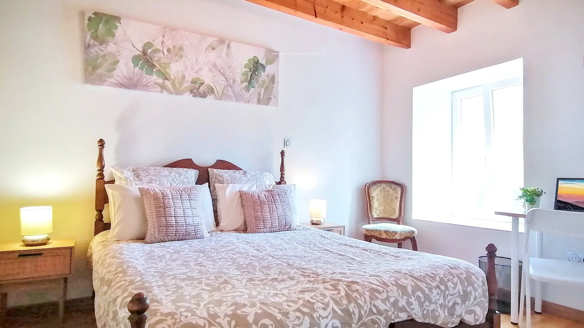 C'est une chambre lumineuse aux couleurs douces (blanc, vert pastel, beige). Le plafond comporte des poutres, les murs sont blancs. Le lit deux personnes est aussi en bois ainsi que la tête de lit. De chaque côté, on trouve des tables de nuit en bois avec une lampe de chevet, ainsi qu'un fauteuil près de la fenêtre et un bureau de l'autre côté avec écran et poubelle à droite du bureau. Les deux pans de murs sont décorés par des tableaux avec des  motifs de végétaux.