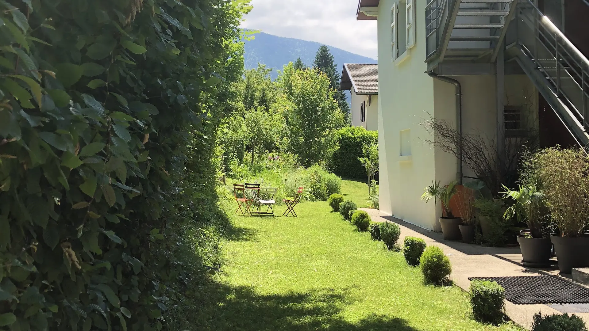 Cette maison de style thermal présente une façade avec des murs peints d'une couleur claire et un toit pentu. De grandes fenêtres laissent entrer la lumière naturelle. Un jardin verdoyant, parsemé d'arbres et d'arbustes, entoure la maison.