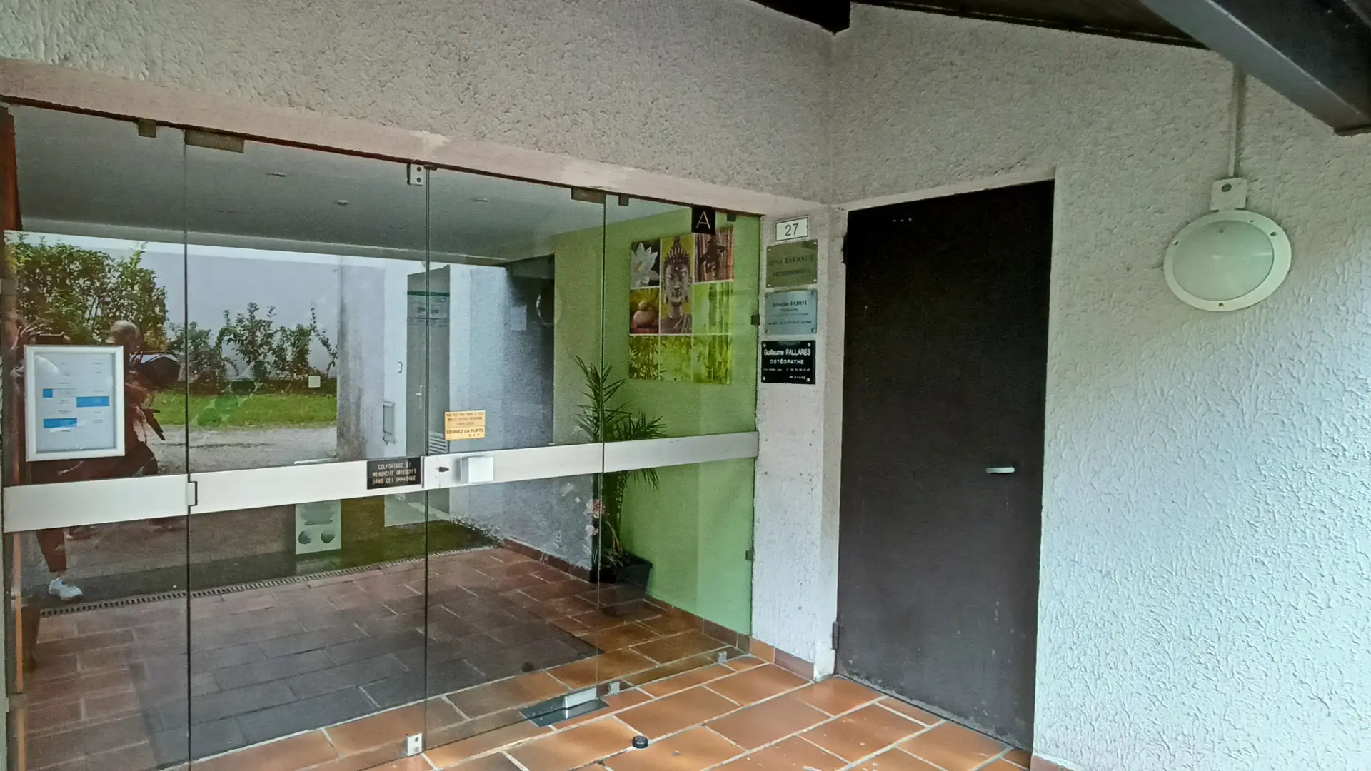 C'est l'entrée d'un bâtiment avec une porte vitrée à gauche, une porte d'entrée noire à droite et un mur en crépi, où l'on trouve des carrelages au sol et des affiches sur le mur près de la porte vitrée.