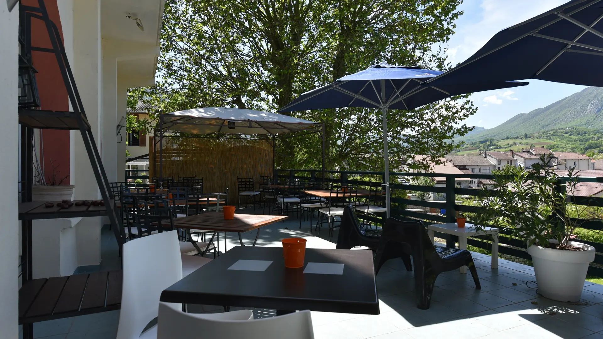 Une pergola et deux grands parasols abritent des tables et des chaises sur une terrasse  spacieuse, le tout baigné de soleil et entouré d’une végétation luxuriante