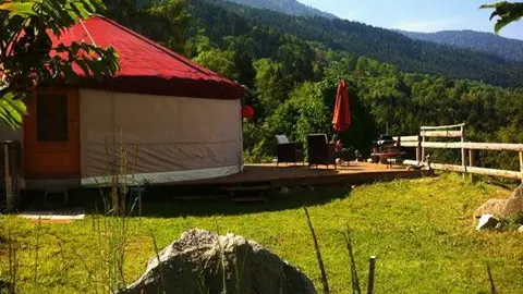 Une yourte traditionnelle, avec son toit en forme de dôme rouge et ses parois beiges, est nichée dans un cadre naturel magnifique. Une terrasse en bois avec des chaises longues invite à la détente.