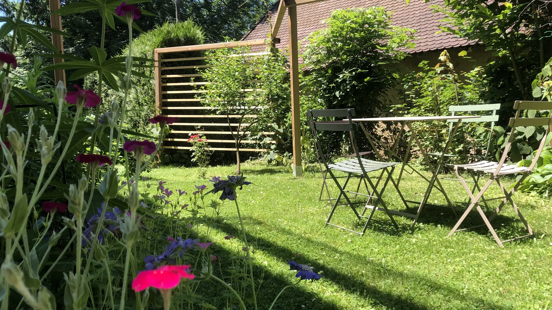 Ce jardin est un espace extérieur aménagé avec une pelouse verdoyante. Un salon de jardin en métal est disposé au centre. Des plantes et des arbustes, dont certaines sont fleuries, bordent le jardin et créent des massifs colorés.