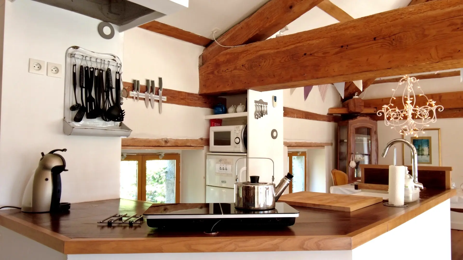 La cuisine se trouve entre la salle à manger et le salon de la mezzanine. Une petite cuisine américaine avec le nécessaire pour cuisiner. Le bar en bois est de forme octogonale ouvert avec un évier, une table à induction et sa bouilloire et contre le mur, une machine à café. On aperçoit des ustensiles de cuisine accroché à un support ancien détourné. A droite, une niche avec une étagère comprenant trois théières. En dessous, il y a un micro-onde et une réfrigérateur (frigo et une partie congélation). Au dessus de la fenêtre, on trouve un support magnétique pour ranger les couteaux de cuisine. La luminosité de la mezzanine est apportée par la lumière traversante venant des fenêtres.