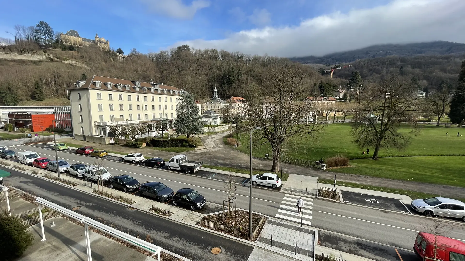 Une scène paisible de ville et de nature se dévoile, avec une rue animée contrastant avec la tranquillité du parc et de la colline du Château d'Uriage