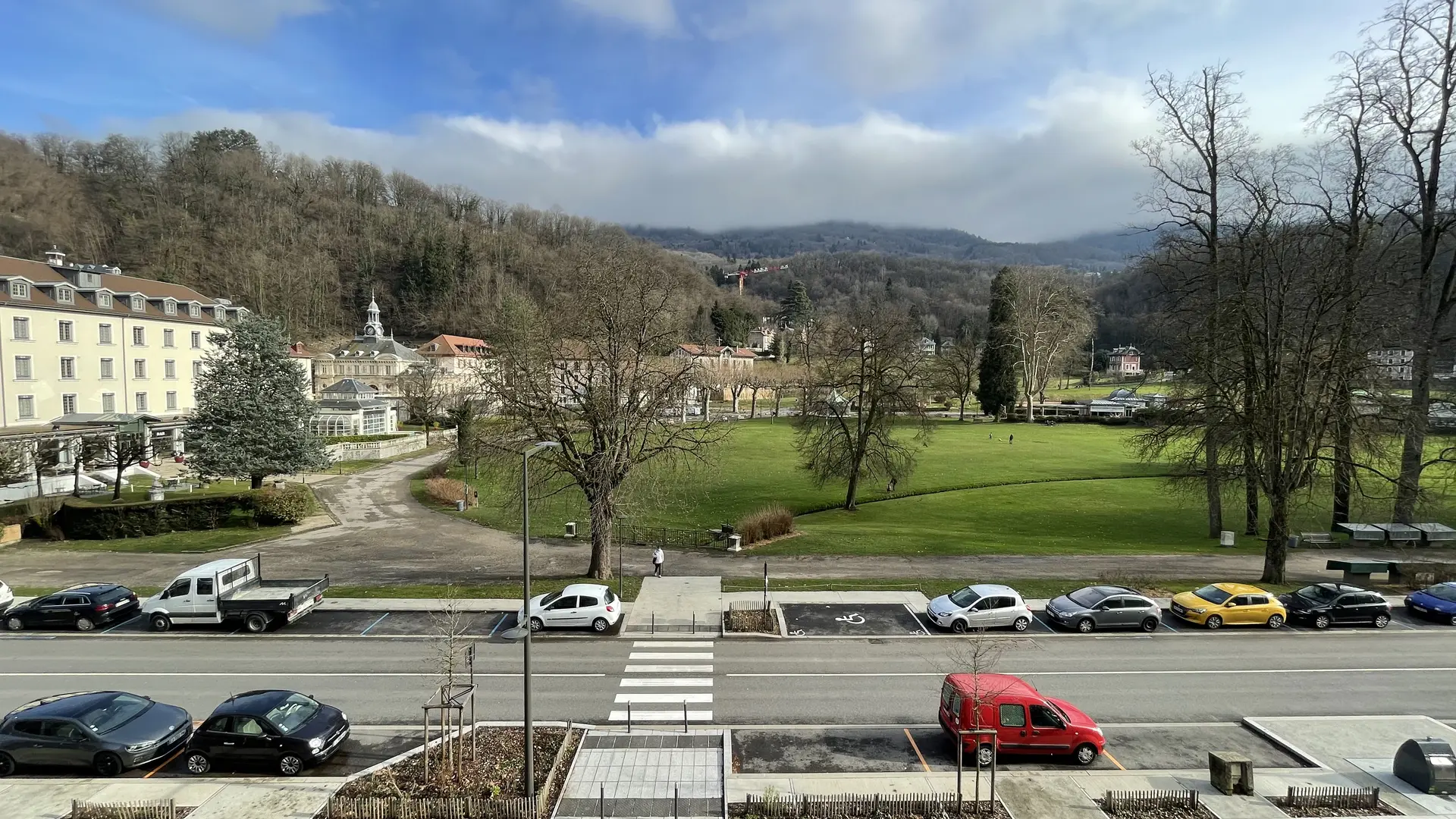 Une vue sur une rue bordée d'arbres, avec des voitures garées des deux côtés, donnant sur le Parc et les thermes d'Uriage et une colline boisée en arrière-plan.