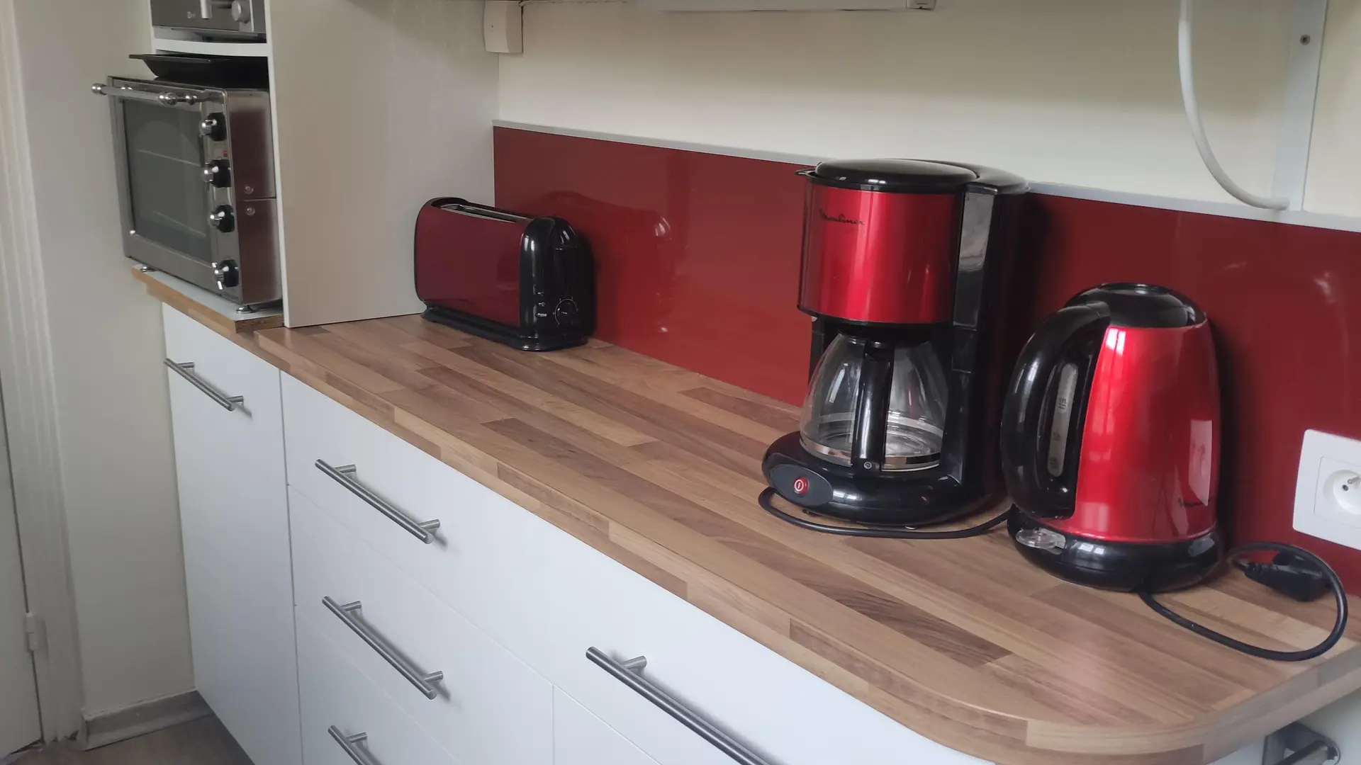 Une cuisine moderne avec des meubles bas blancs à tiroirs, un plan de travail effet bois, une crédence rouge et des étagères murales blanches.