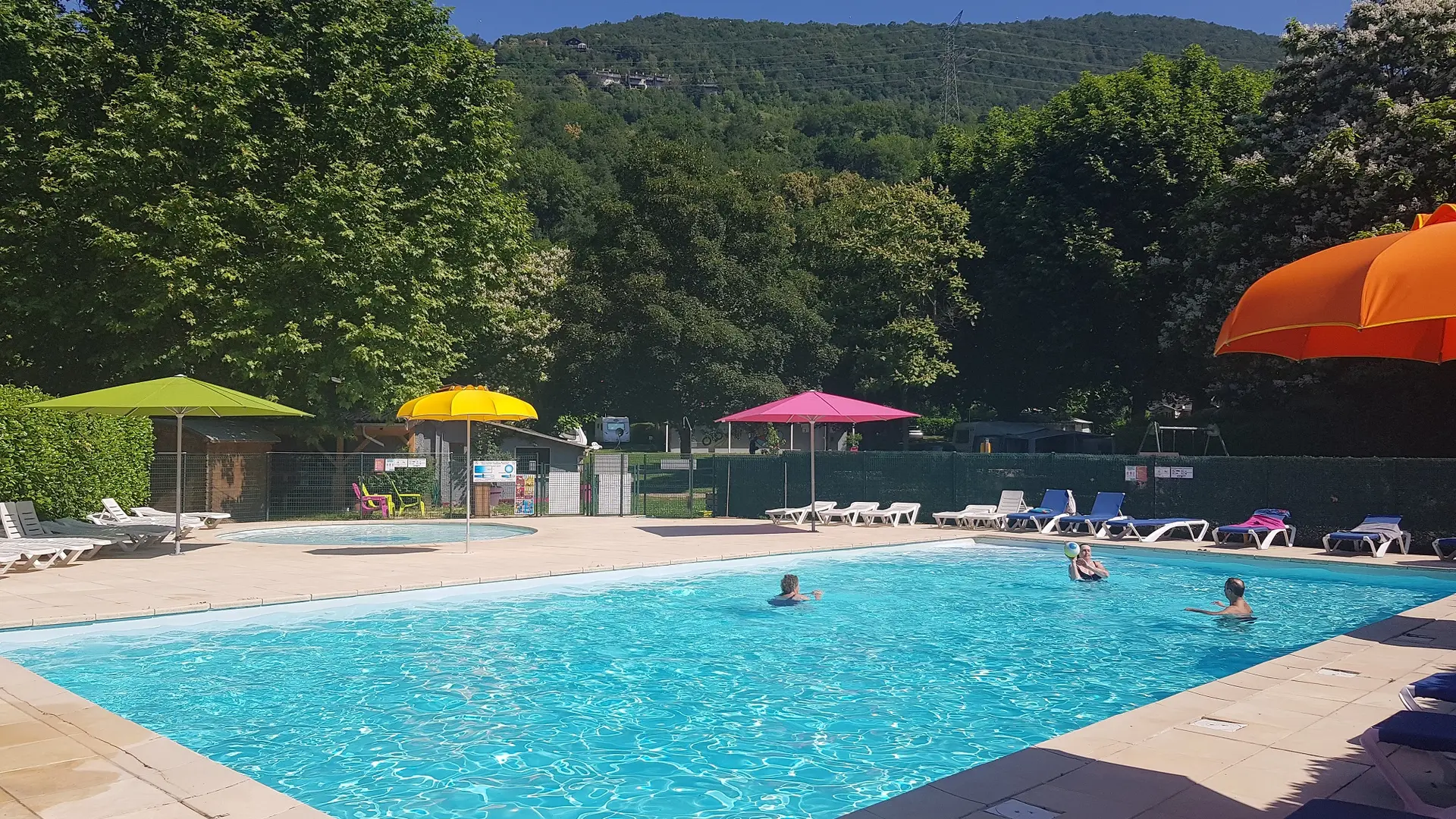 Une piscine rectangulaire est entourée d'une terrasse ensoleillée. Des parasols colorés sont plantés autour de la piscine, offrant de l'ombre aux nageurs. Des chaises longues sont disposées sur la terrasse. On distingue une pataugeoire ronde en arrière-plan.