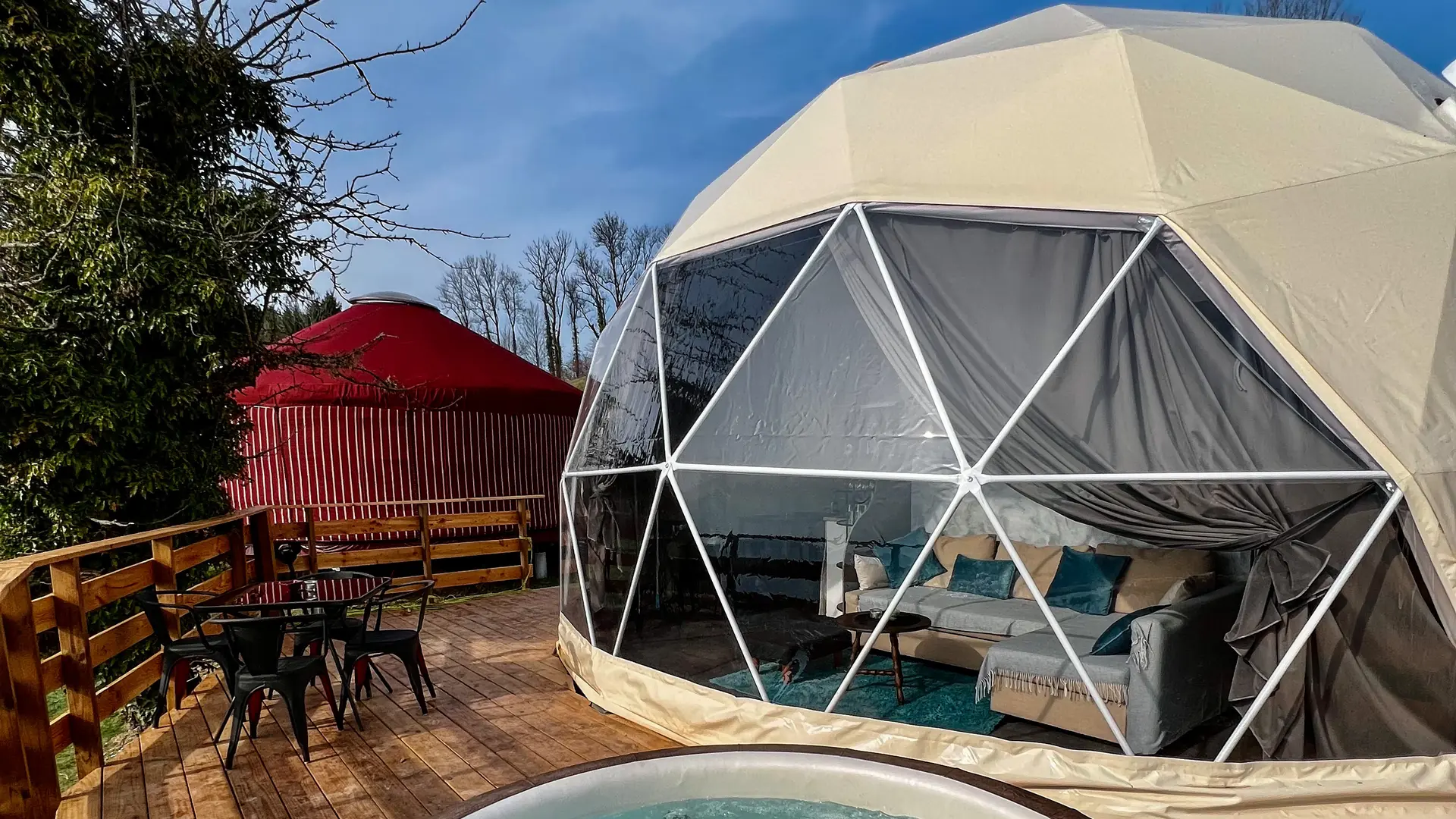 Un dôme géodésique blanc aux parois transparentes, situé sur une terrasse en bois avec un salon de jardin et un spa rond rempli d'eau. À proximité, on peut distinguer une yourte de couleur rouge.