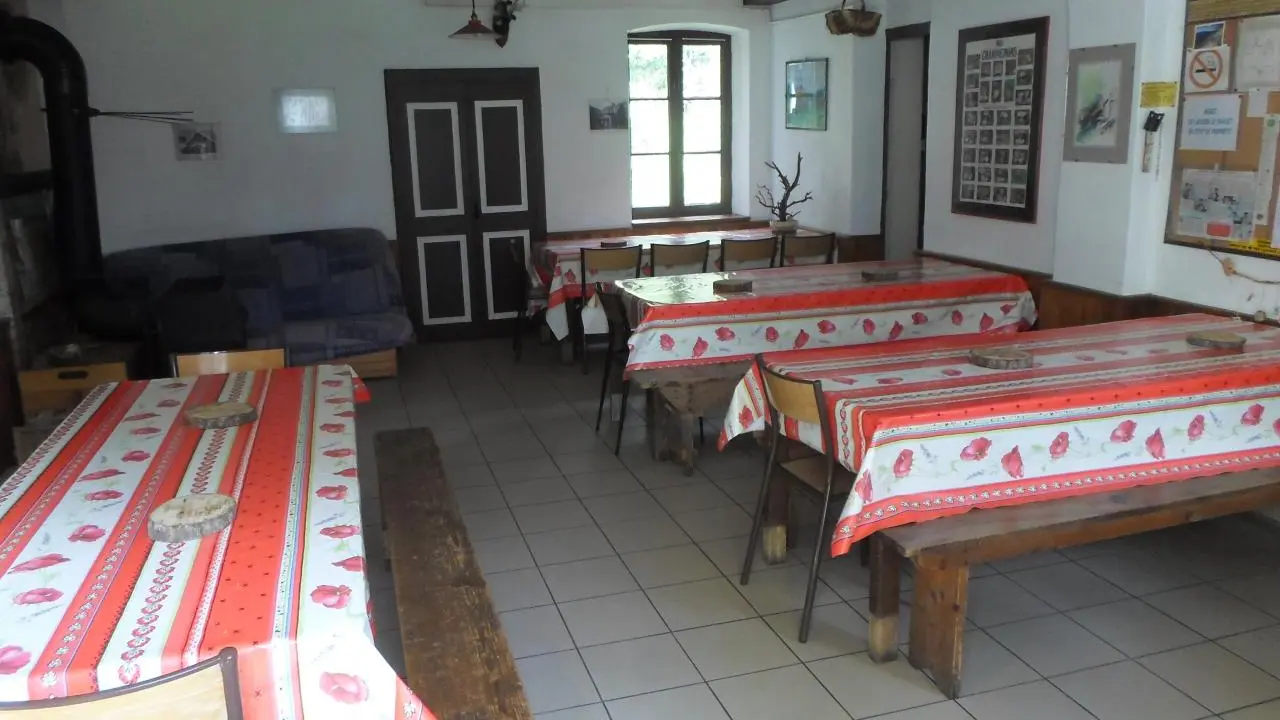 Il s'agit d'une grande pièce rectangulaire avec un plafond en bois apparent. Le sol est en carrelage. Plusieurs tables en bois, recouvertes de nappes à carreaux rouges et blancs, sont disposées dans la pièce. Des chaises et des bancs en bois complètent l'aménagement.