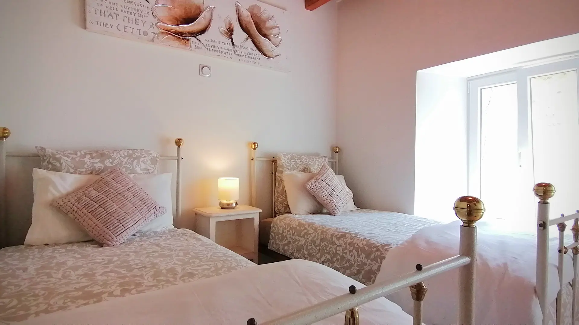 Même atmosphère que la chambre avec le lit pour 2 personnes. La chambre comporte deux lits en métal blanc et doré à une personne et entre une table de chevet avec un luminaire. Une fenêtre illumine la pièce. Le plafond est en bois, les murs blancs agrémenté d'un tableau beige et doré avec l'impression de végétaux et d'inscription en anglais.