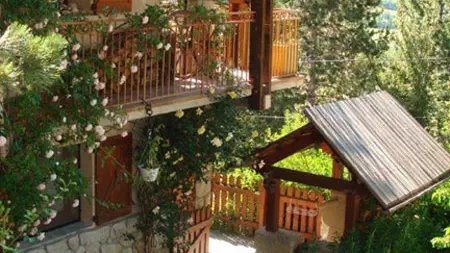 Vue de côté d'une maison sur 3 étages  en bois et pierre avec balcons métalliques, petite cour intérieur, abri de jardin, paysage printanier