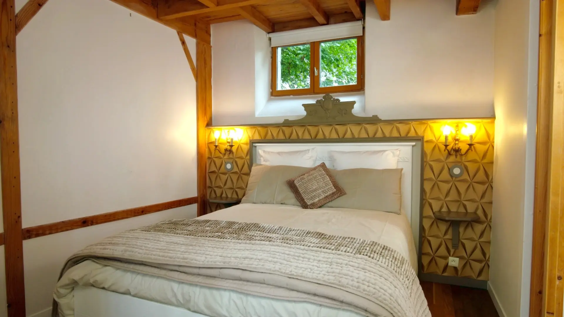 Nous sommes dans la première chambre, en face d'un lit deux place avec une tête de lit à motif géométrique vintage jaune et gris avec les appliques et tables de chevet intégrés. Au-dessus, une petite fenêtre laisse rentrer une douce lumière. Le bois est aussi présent dans cette pièce avec des galandages à colombages. Le linge de lit s'accorde avec la tête de lit. Le sol est en plancher
