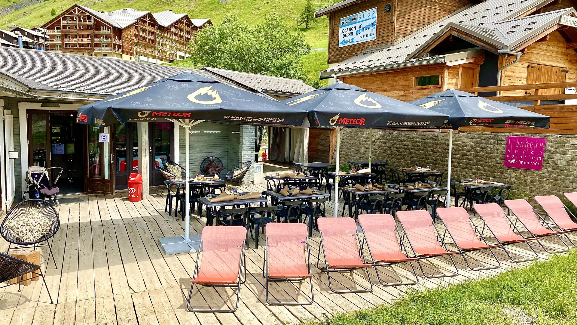 Terrasse de l'établissement en été, tables, chaises, parasols, chaises longues, au milieu de la station et de végétation