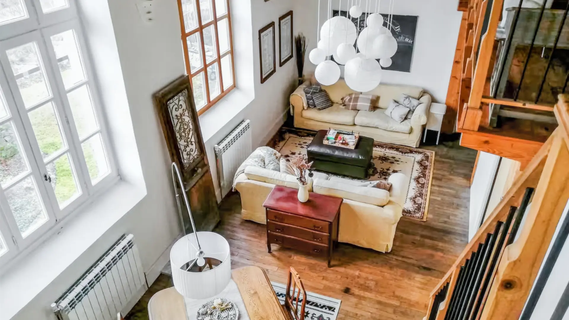 Une belle pièce de vie très lumineuse avec ses trois grandes fenêtres. Elle est toute en longueur avec plusieurs postes de loisirs,. Deux salons et entre les deux une table pour faire des jeux avec un lampadaire à arc. On trouve un poêle à bois au fond de la salle avec à gauche du poêle, un guéridon bas et une lampe avec un serviteur contenant le bois et à droite une armoire vintage peinte en blanc. A côté du canapé, on retrouve un autre guéridon et une lampe avec au-dessus un grand miroir sur le mur. Au premier plan, deux autres canapés et une table basse. en décoration, une veille porte à côté du canapé. Le lustre de la pièce est composé de plusieurs lanternes en papier blanc. Sur la gauche, un escalier en bois permet d'accéder à la mezzanine.
