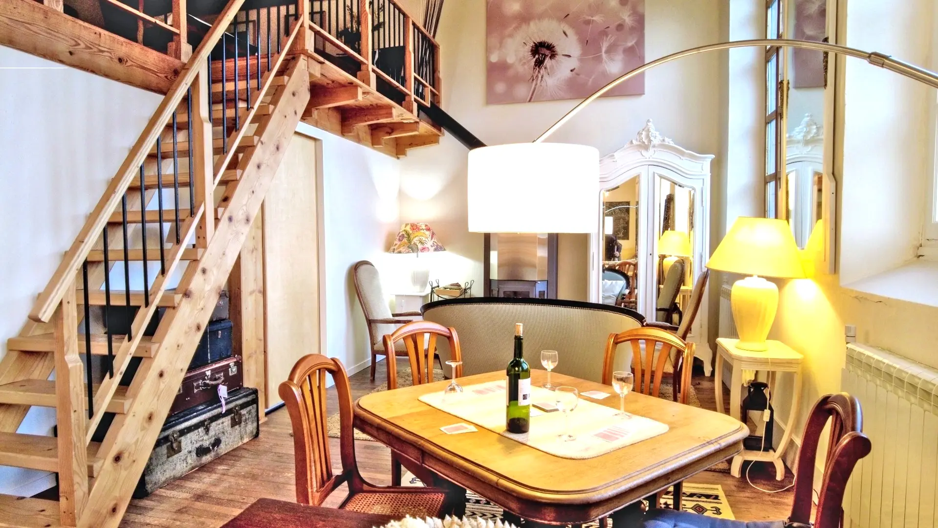 Une belle pièce de vie très lumineuse avec ses trois grandes fenêtres. Elle est toute en longueur avec plusieurs postes de loisirs,. Deux salons et entre les deux une table pour faire des jeux avec un lampadaire à arc. On trouve un poêle à bois au fond de la salle avec à gauche du poêle, un guéridon bas et une lampe avec un serviteur contenant le bois et à droite une armoire vintage peinte en blanc. A côté du canapé, on retrouve un autre guéridon et une lampe avec au-dessus un grand miroir sur le mur. Au premier plan, deux autres canapés et une table basse. en décoration, une veille porte à côté du canapé. Le lustre de la pièce est composé de plusieurs lanternes en papier blanc. Sur la gauche, un escalier en bois permet d'accéder à la mezzanine.