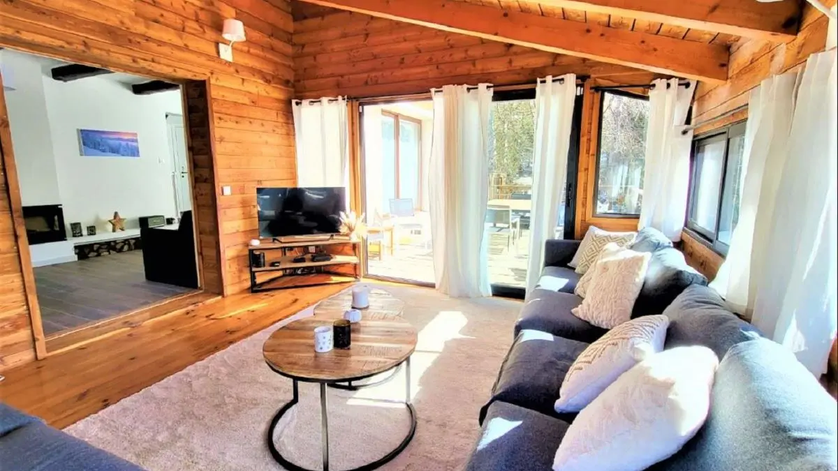 Séjour lumineux dans une véranda en bois, avec canapé, tapis, table basse, TV, porte-fenêtre, jonction avec un salon intérieur carrelé