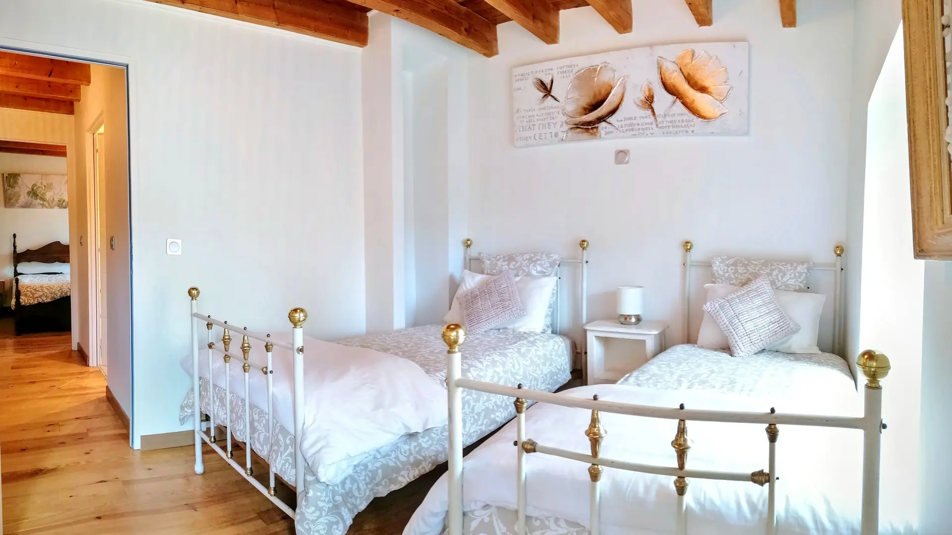 Même atmosphère que la chambre avec le lit pour 2 personnes. La chambre comporte deux lits en métal blanc et doré à une personne et entre une table de chevet avec un luminaire. Une fenêtre illumine la pièce. Le plafond est en bois, les murs blancs agrémenté d'un tableau beige et doré avec l'impression de végétaux et d'inscription en anglais.