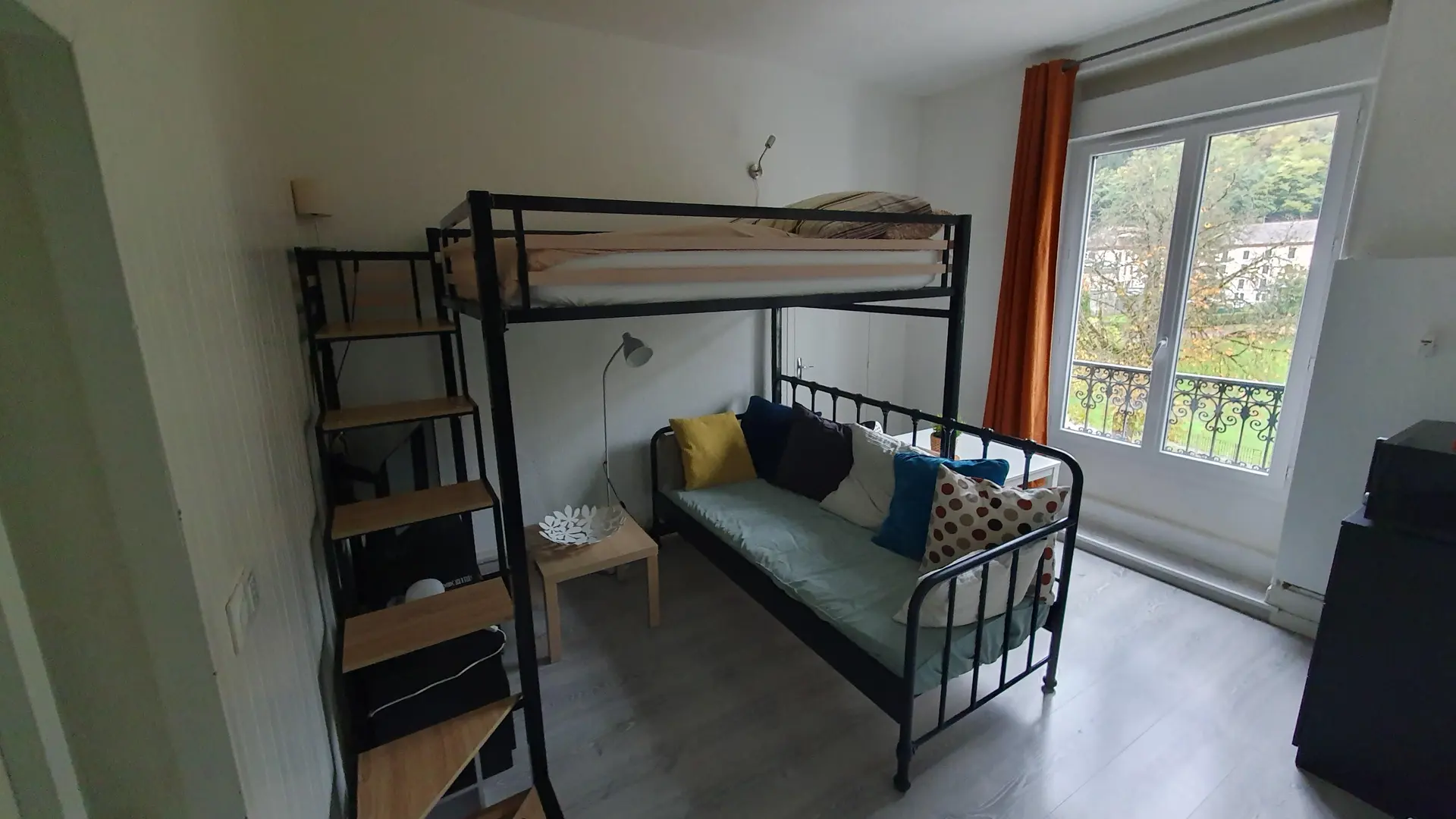 La pièce est un studio de style moderne contenant un lit mezzanine en métal noir avec un canapé-lit en dessous décoré de coussins colorés, une étagère latérale servant d'escalier, et une grande fenêtre avec un rideau orange donnant sur le parc thermal.