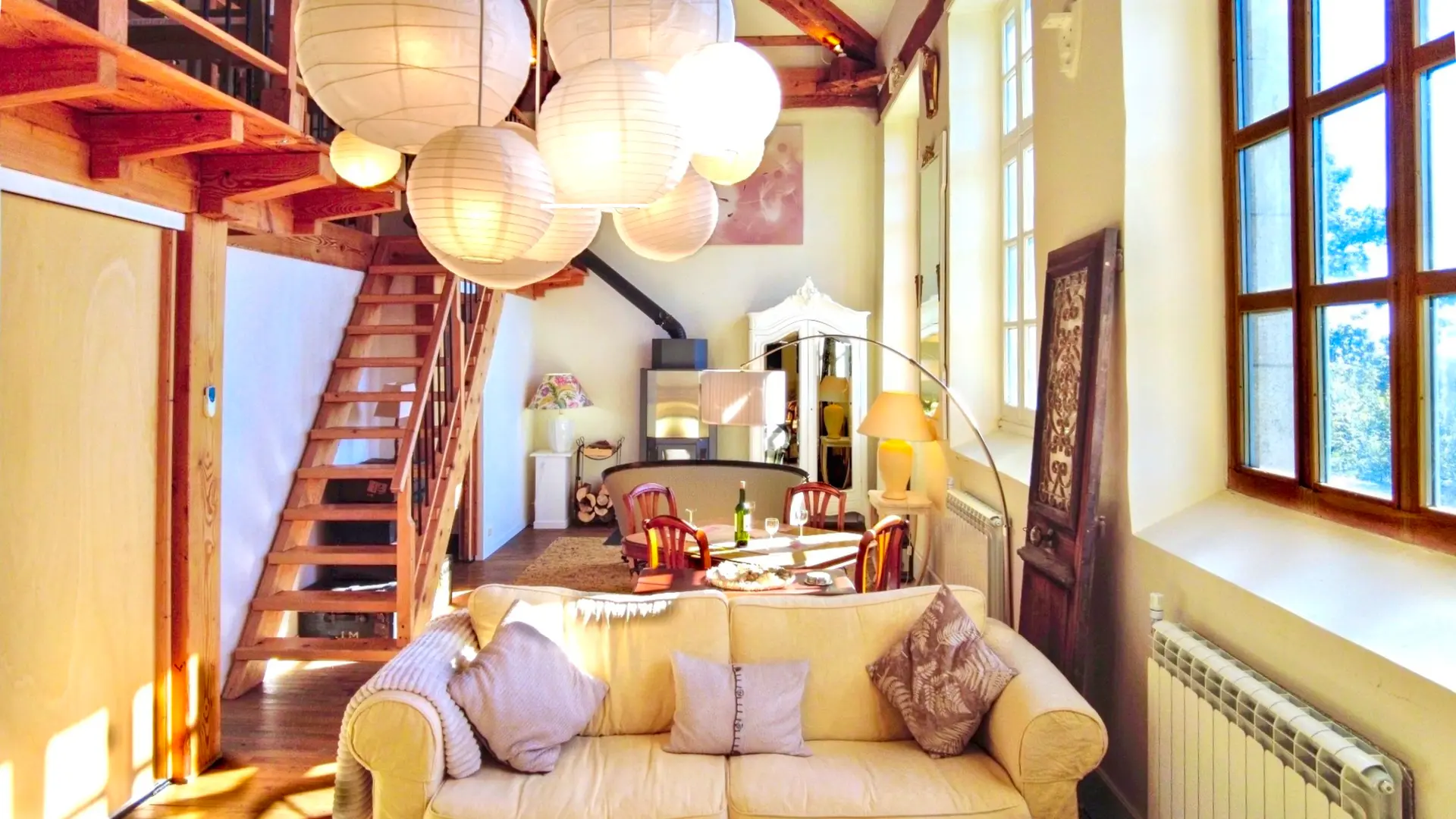 Une belle pièce de vie très lumineuse avec ses trois grandes fenêtres. Elle est toute en longueur avec plusieurs postes de loisirs,. Deux salons et entre les deux une table pour faire des jeux avec un lampadaire à arc. On trouve un poêle à bois au fond de la salle avec à gauche du poêle, un guéridon bas et une lampe avec un serviteur contenant le bois et à droite une armoire vintage peinte en blanc. A côté du canapé, on retrouve un autre guéridon et une lampe avec au-dessus un grand miroir sur le mur. Au premier plan, deux autres canapés et une table basse. en décoration, une veille porte à côté du canapé. Le lustre de la pièce est composé de plusieurs lanternes en papier blanc. Sur la gauche, un escalier en bois permet d'accéder à la mezzanine.