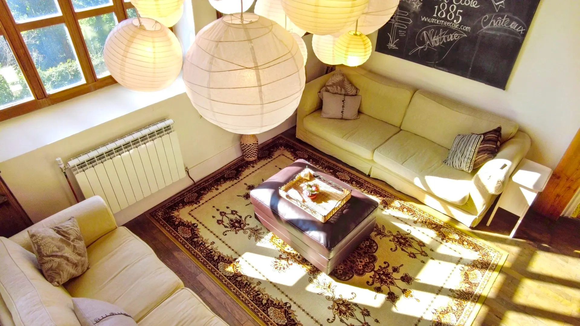 Une belle pièce de vie très lumineuse avec ses trois grandes fenêtres. Elle est toute en longueur avec plusieurs postes de loisirs,. Deux salons et entre les deux une table pour faire des jeux avec un lampadaire à arc. On trouve un poêle à bois au fond de la salle avec à gauche du poêle, un guéridon bas et une lampe avec un serviteur contenant le bois et à droite une armoire vintage peinte en blanc. A côté du canapé, on retrouve un autre guéridon et une lampe avec au-dessus un grand miroir sur le mur. Au premier plan, deux autres canapés et une table basse. en décoration, une veille porte à côté du canapé. Le lustre de la pièce est composé de plusieurs lanternes en papier blanc. Sur la gauche, un escalier en bois permet d'accéder à la mezzanine.