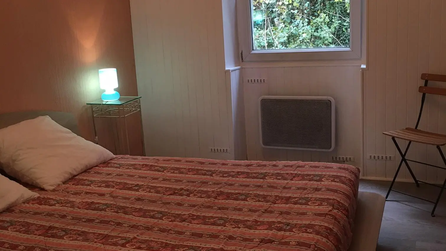 C'est une chambre avec un lit double au centre. Il y a une fenêtre à droite du lit qui donne sur de la végétation. À côté du lit, il y a une petite table de nuit avec une lampe. Dans le coin, il y a une chaise.