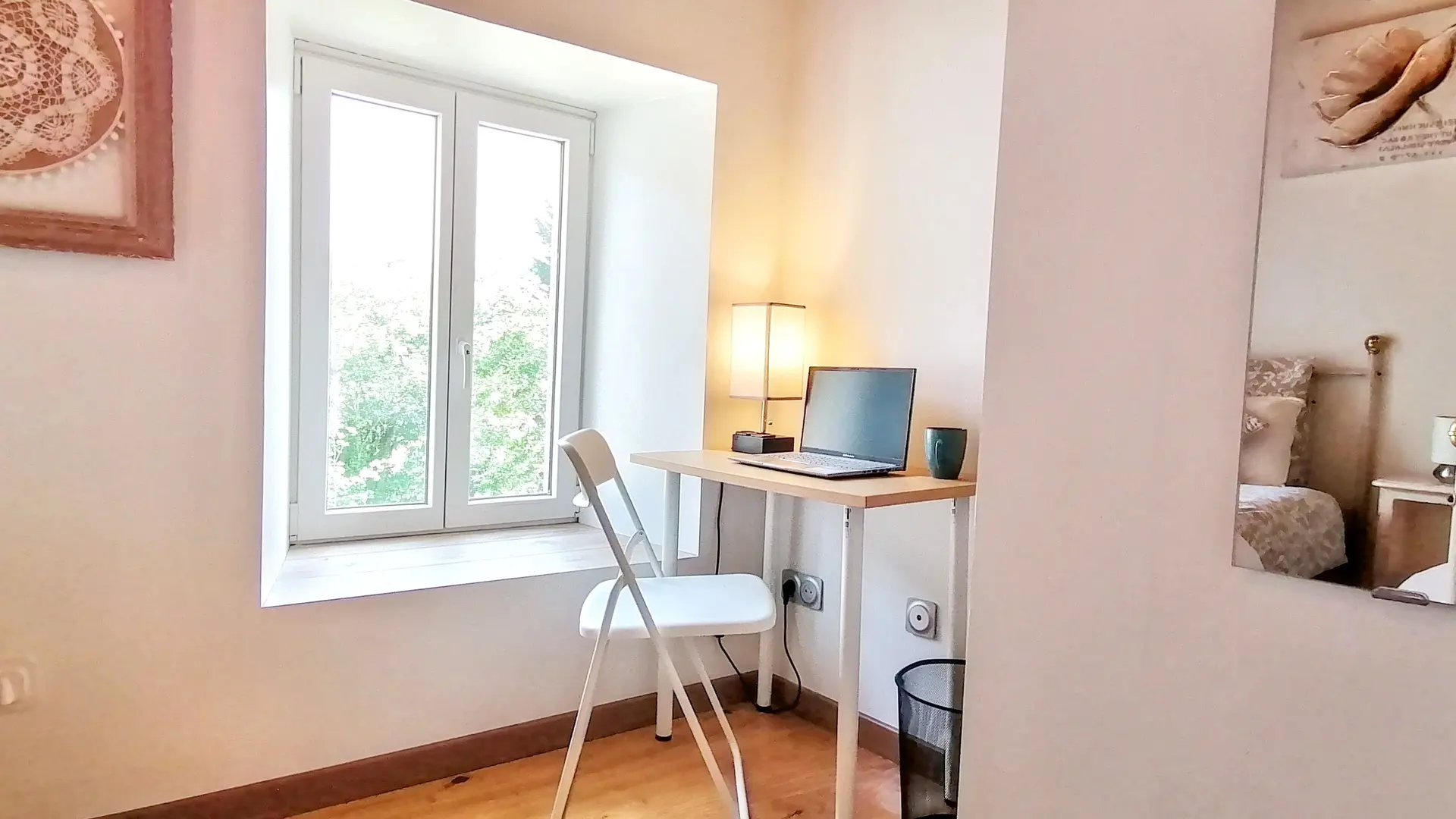 Un coin bureau simple et lumineux dans une chambre à coucher. Au centre, une fenêtre à double battant laisse entrer la lumière naturelle et offre une vue sur la verdure extérieure. Sous la fenêtre, un petit bureau rectangulaire en bois clair avec des pieds métalliques blancs est installé. Un ordinateur portable et une lampe de bureau sont posés sur le bureau, ainsi qu'une tasse. Une chaise pliante blanche est placée devant le bureau.  Le mur est blanc et à l'extrême gauche, un tableau encadré d'une broderie de couleur crème est accroché. À droite, un grand miroir mural reflète la pièce, révélant une partie d'un lit avec une tête de lit en métal et une table de chevet avec une lampe. Le sol est un plancher en bois clair.