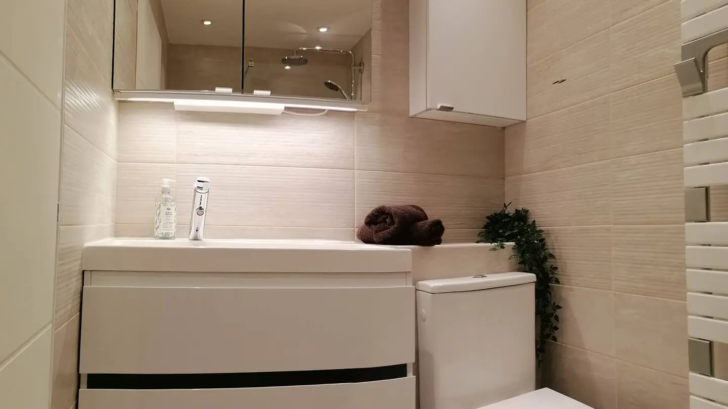 C'est une salle de douche avec un wc, un lavabo une vasque et un meuble de rangement en-dessous et au-dessus. La salle de douche est carrelé de haut en bas. A droite, il y a un sèche serviette et au-dessus des wc se trouve aussi un meuble de rangement. Les couleurs sont douce entre le blanc et le beige.