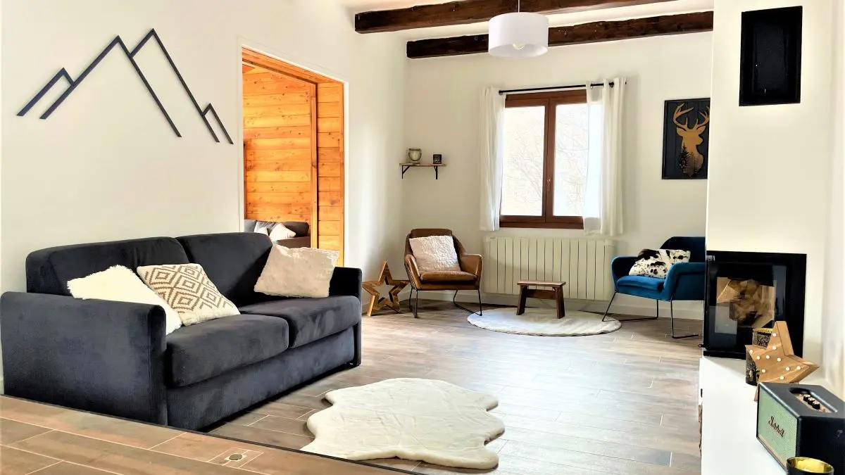 Grand séjour lumineux avec cheminée, canapé, fauteuils, ameublement bois, poutres apparentes, parquet au sol, fenêtre