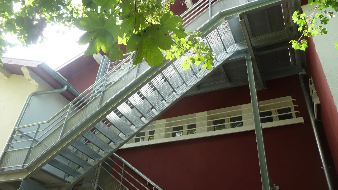 C'est un escalier métallique extérieur avec des marches plates et des rampes solides. Il est adossé à la maison et donne accès à un étage supérieur.