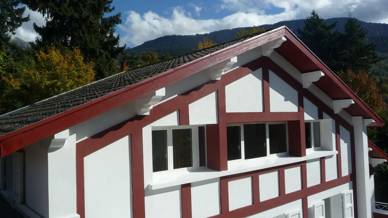 Cette maison de style thermal présente des murs à colombage et un toit pentu recouvert de tuiles rouges. Elle est implantée dans un cadre verdoyant, avec une vue sur les montagnes.